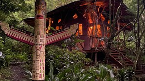 Uma cabana de madeira rústica com telhado de palha, rodeada por vegetação exuberante e um totém decorativo.
