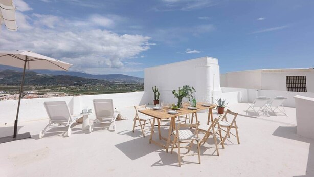 Una terraza en la azotea con una mesa y sillas de madera, una planta de cactus y vistas al paisaje.