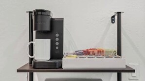 Una cafetera, una taza blanca y una caja de bolígrafos sobre una mesa.