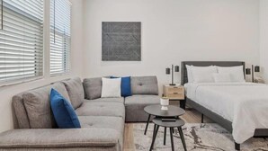 Un dormitorio moderno con un sofá gris, una mesita redonda, una cama con ropa de cama blanca y una obra de arte geométrica en la pared.