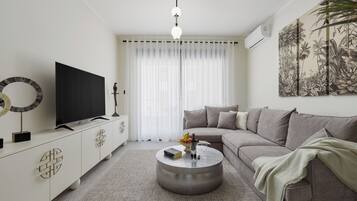 Una sala de estar moderna con un televisor de pantalla plana, una mesa redonda de centro con libros y una botella, un sofá gris y una gran obra de arte en la pared.