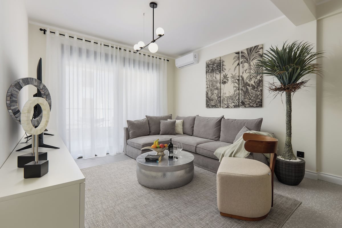 A modern living room with a sectional sofa, a round coffee table with fruit and a bottle, a side table with a decorative mirror, a potted plant, and a wall-mounted air conditioner.