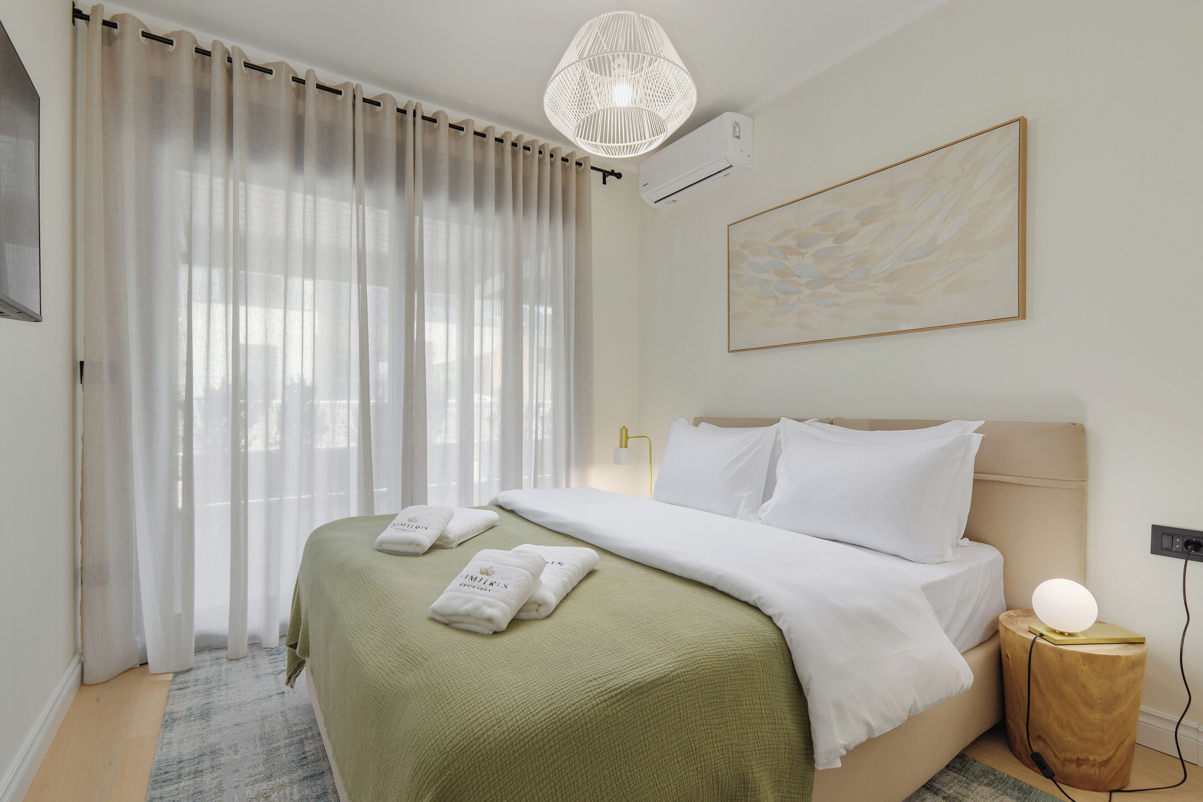 Een netjes opgemaakt bed met een groene dekbedovertrek, witte kussens en twee handdoeken. Er staat een bedlampje, een wandairconditioner en er hangt een schilderij boven het bed.