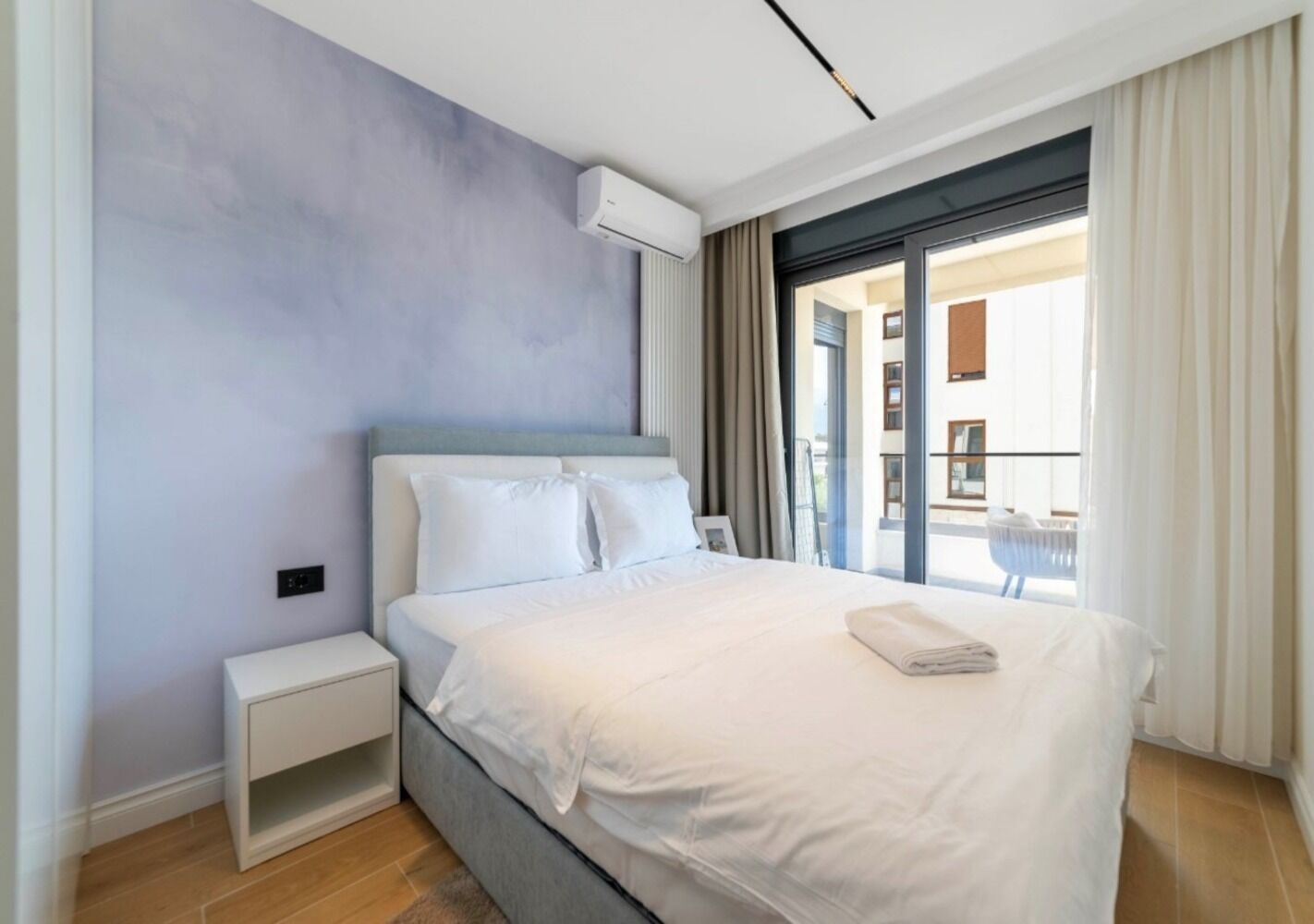 A modern hotel room with a large bed, a bedside table, and a view of the outside balcony.