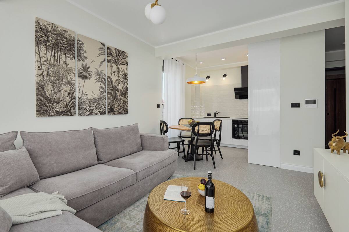 A modern living room with a grey sofa, a round coffee table with a bottle of wine and a glass, and a dining area with a small table and chairs.
