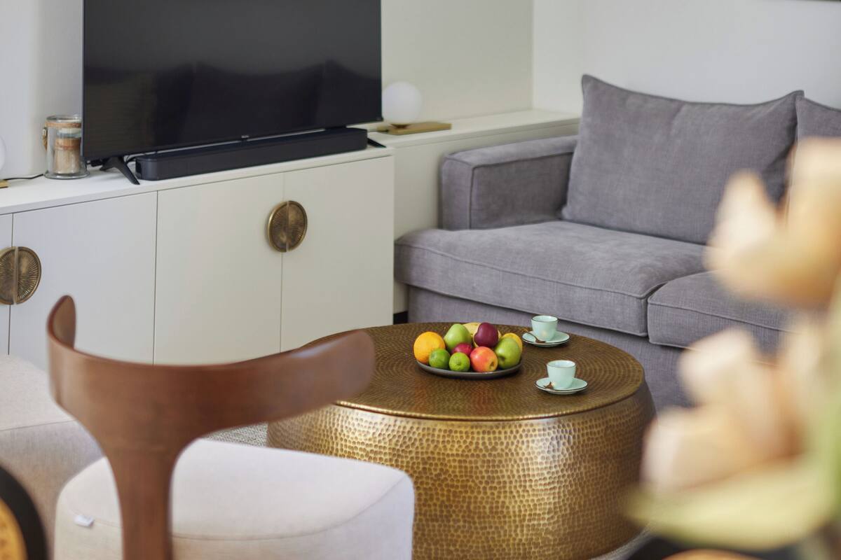 A modern living room with a flat-screen TV, grey sofa, round coffee table with fruit, and a woven lamp.