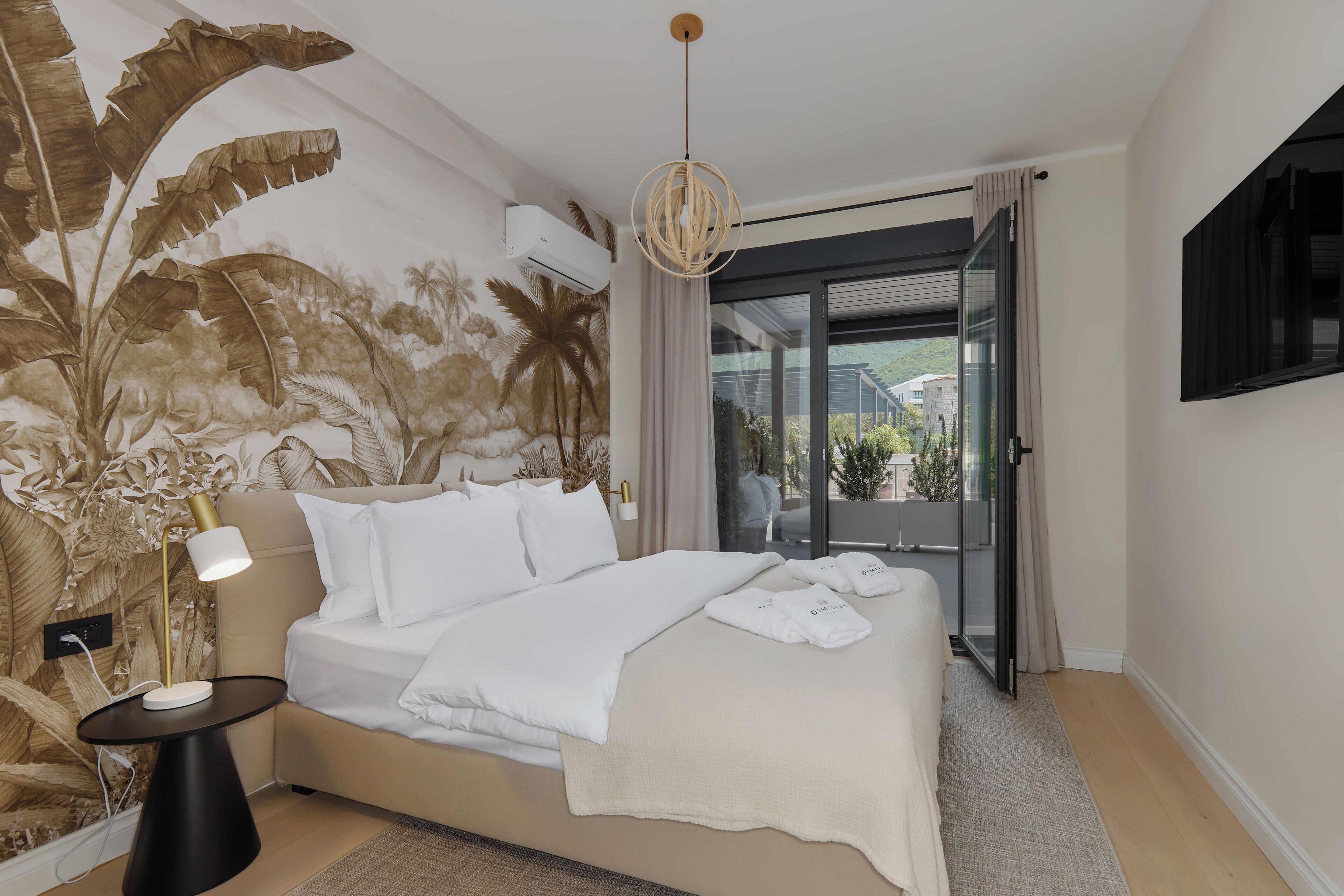Habitación de hotel moderna con una cama grande, una mesita de noche con lámpara, un televisor montado en la pared y vistas al exterior a través de una puerta corrediza de vidrio.