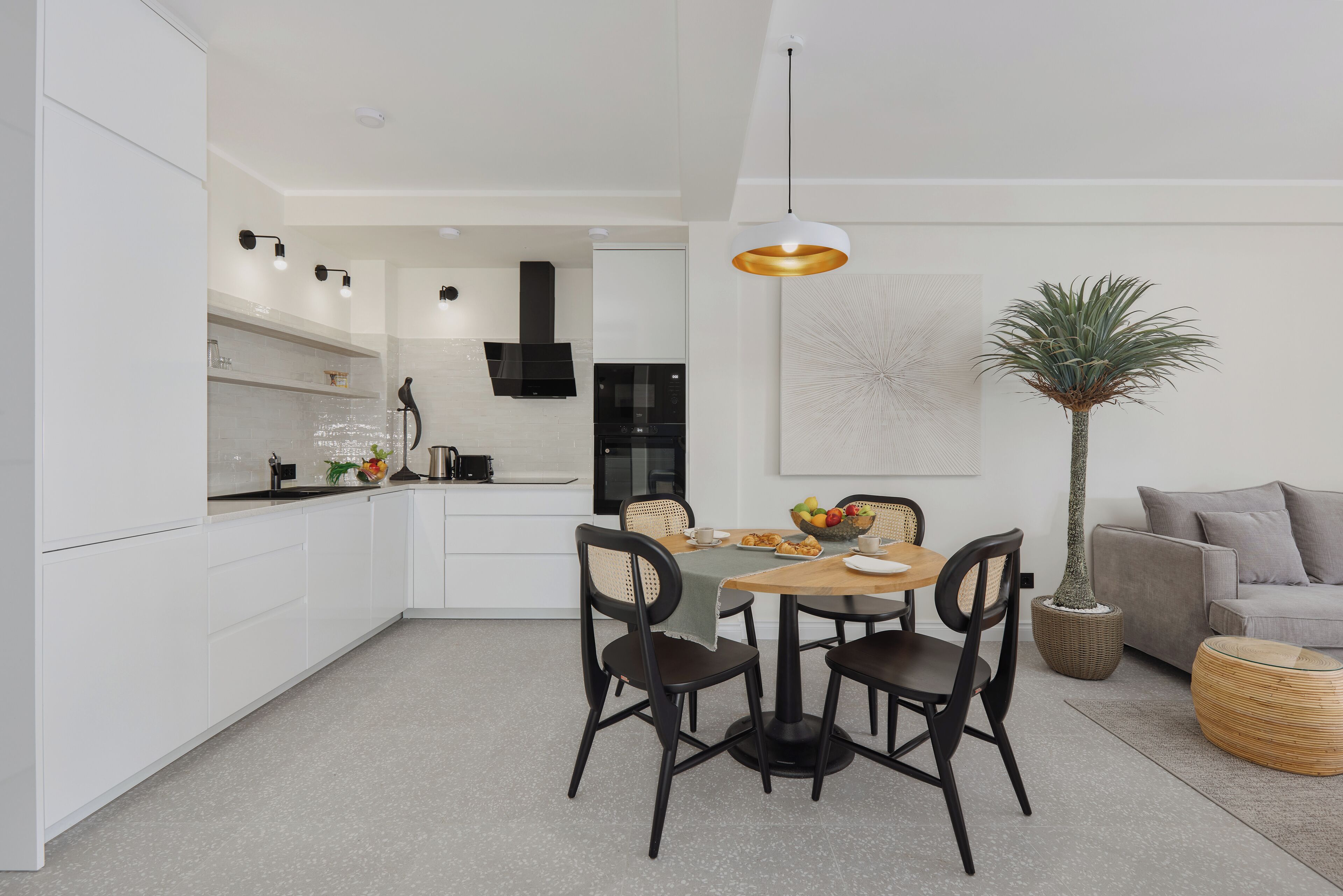 Une cuisine moderne avec des meubles blancs, un coin repas doté d’une table ronde et de chaises, et un canapé gris dans l’espace salon.
