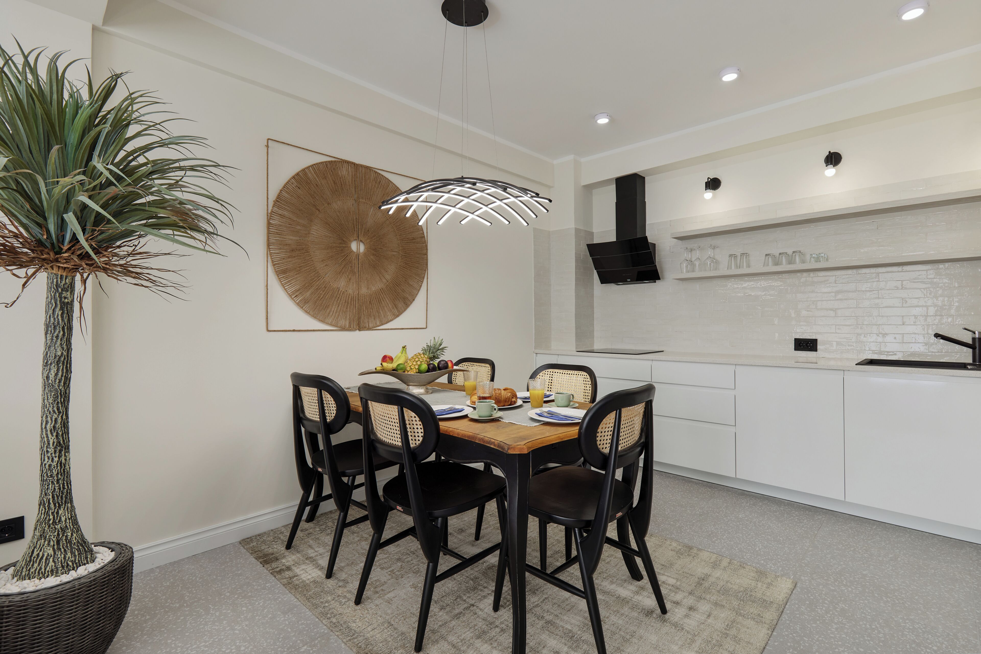 A modern kitchen with white cabinetry, a dining table with four chairs, a large potted plant, and a unique wall art piece.