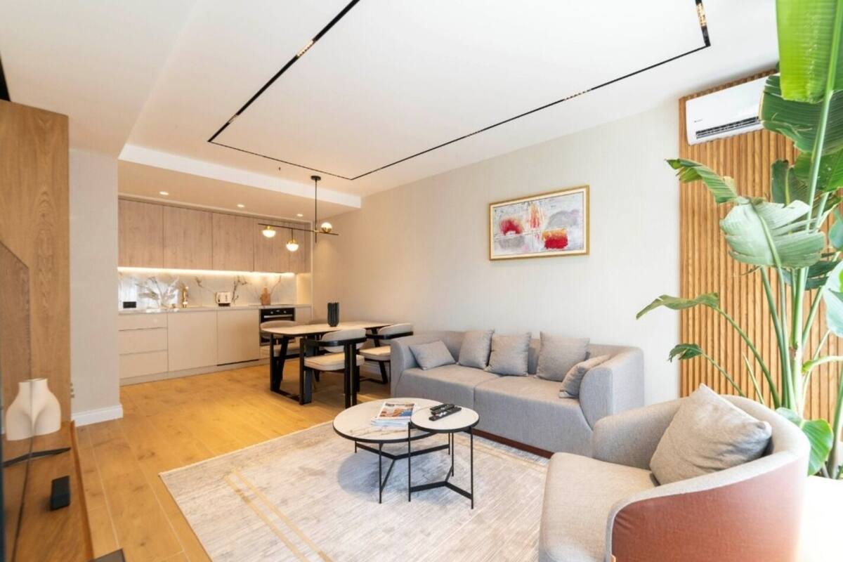 A modern living room with a grey sofa, a coffee table, and a dining area with a table and chairs. There is a kitchen in the background with wooden cabinets and a hanging light fixture. A large plant is visible in the corner.