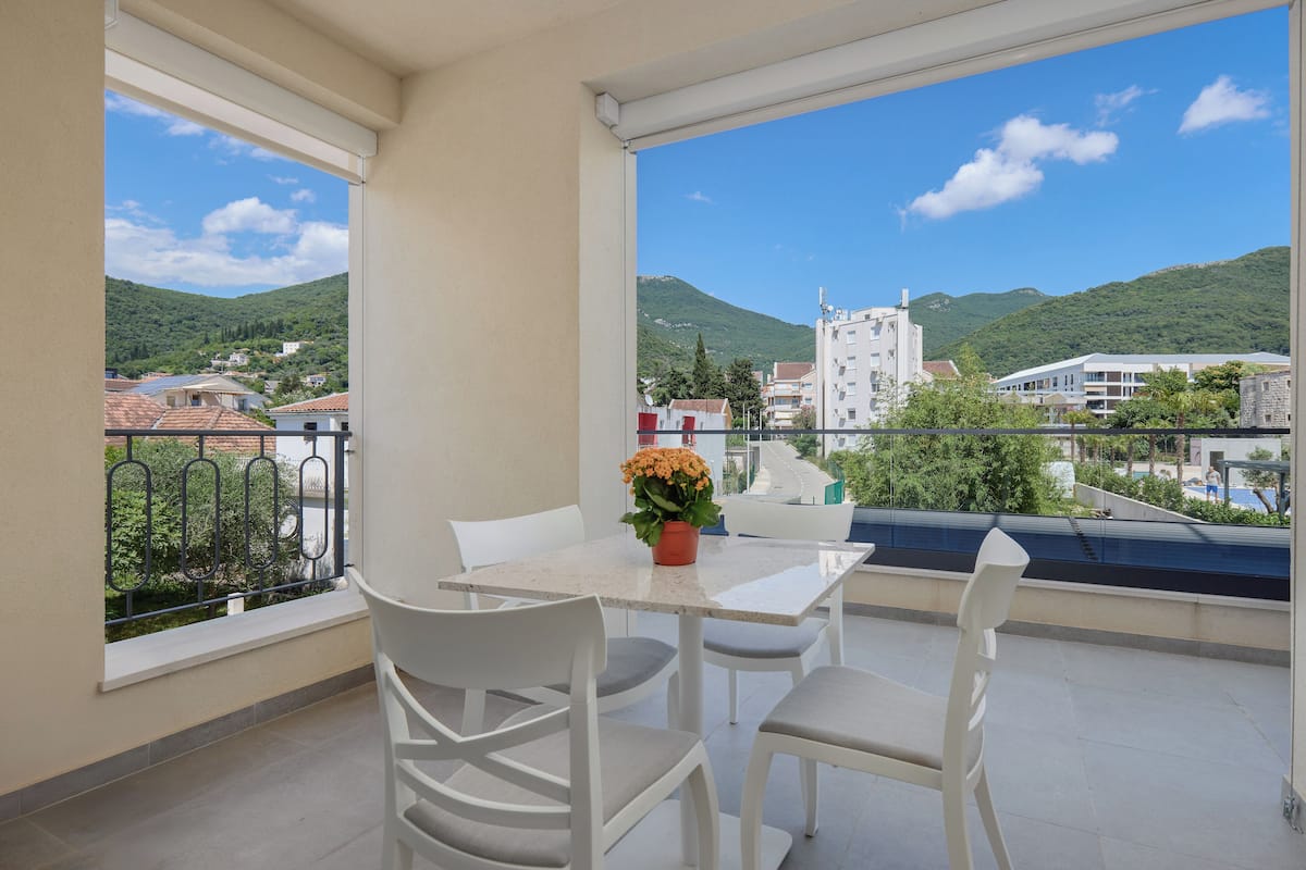 A balcony with a table and chairs, a potted plant, and a view of a town with buildings and hills.