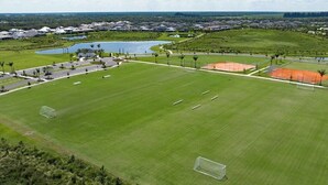En vy från ovan av en stor, välskött sportplan med flera fotbollsmål, omgiven av grönområden och en liten sjö.