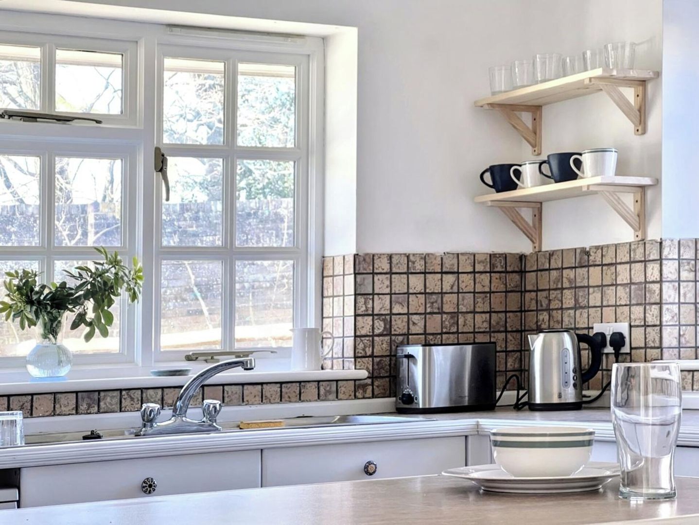 A kitchen with a window, a sink, a kettle, and a shelf with cups.