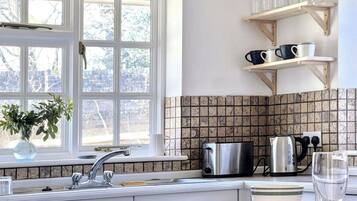 A kitchen with a window, a sink, a kettle, and a shelf with cups.