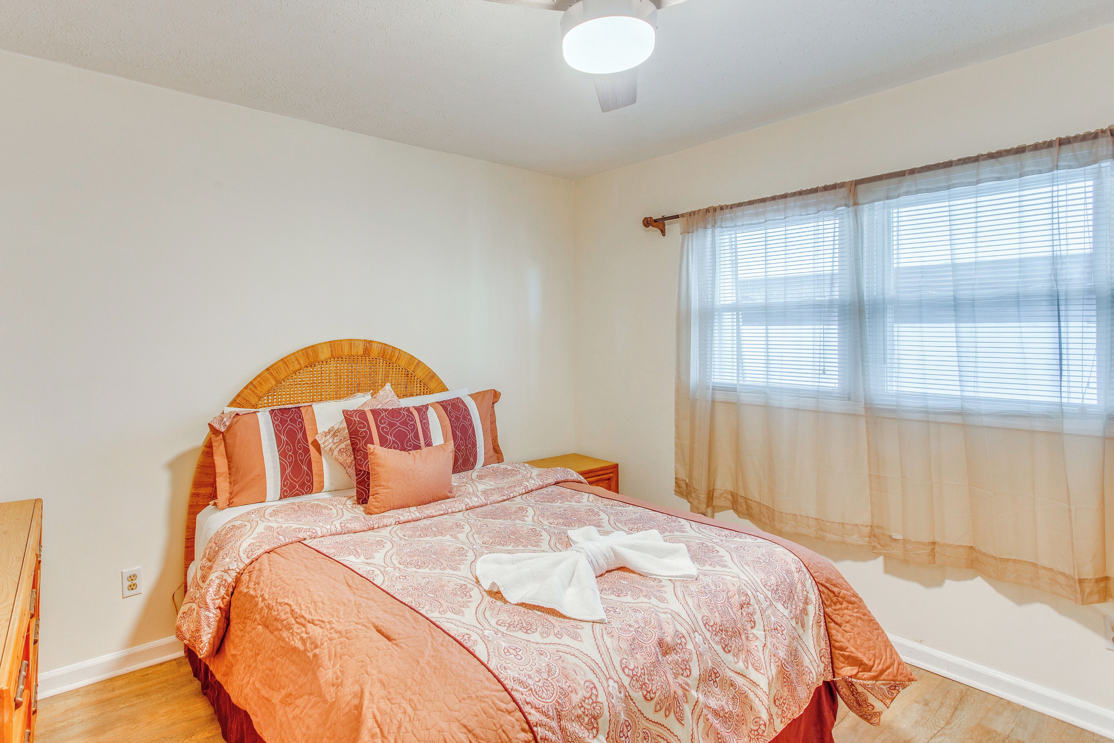 A neatly made bed with a patterned bedspread, a wooden headboard, and a window with sheer curtains.