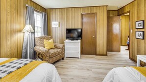 Ein Zimmer mit Holzwandverkleidung, einem Fernseher auf einer weißen Kommode, einem gemusterten Sessel und zwei Betten mit weißer Bettwäsche.