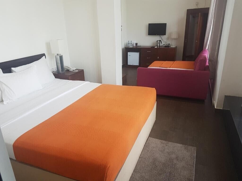A hotel room with a bed, a desk with a lamp, a television, a small kitchenette, and a pink chair.