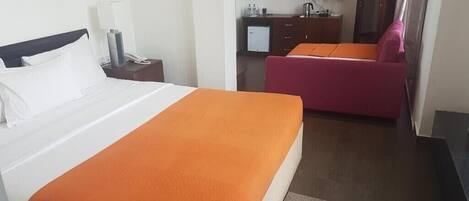A hotel room with a bed, a desk with a lamp, a television, a small kitchenette, and a pink chair.