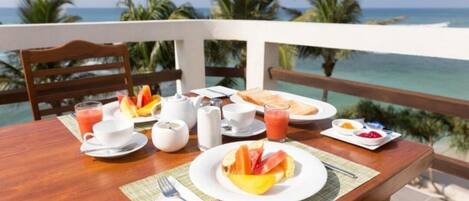 A breakfast table with plates of fruit, juice, and coffee, set against a view of the ocean and palm trees.