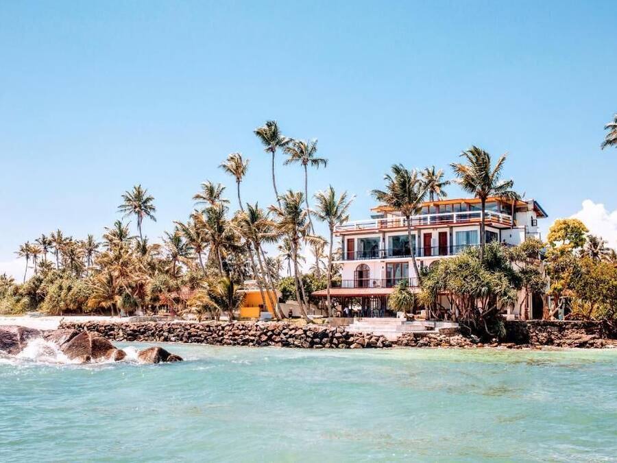 Una propiedad frente a la playa con un edificio grande de varios pisos, palmeras y una costa rocosa.