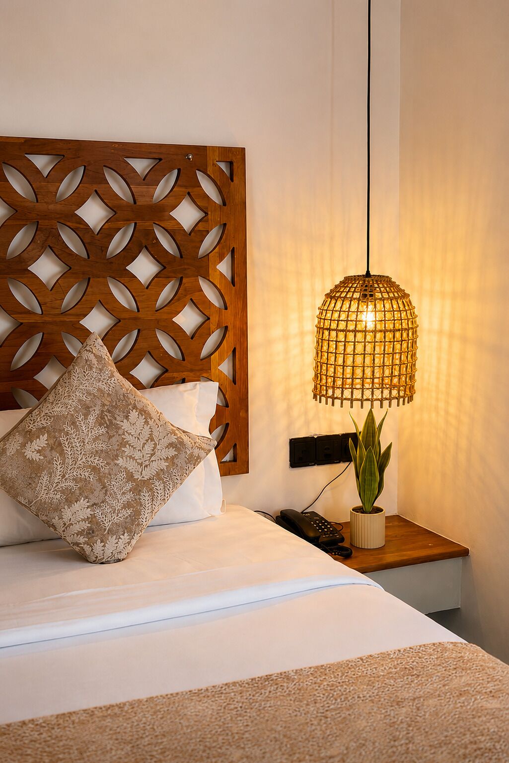 Una cama con cabecera de madera, un cojín estampado, una lámpara colgante, una planta en la mesita de noche y un teléfono en la mesita de noche.