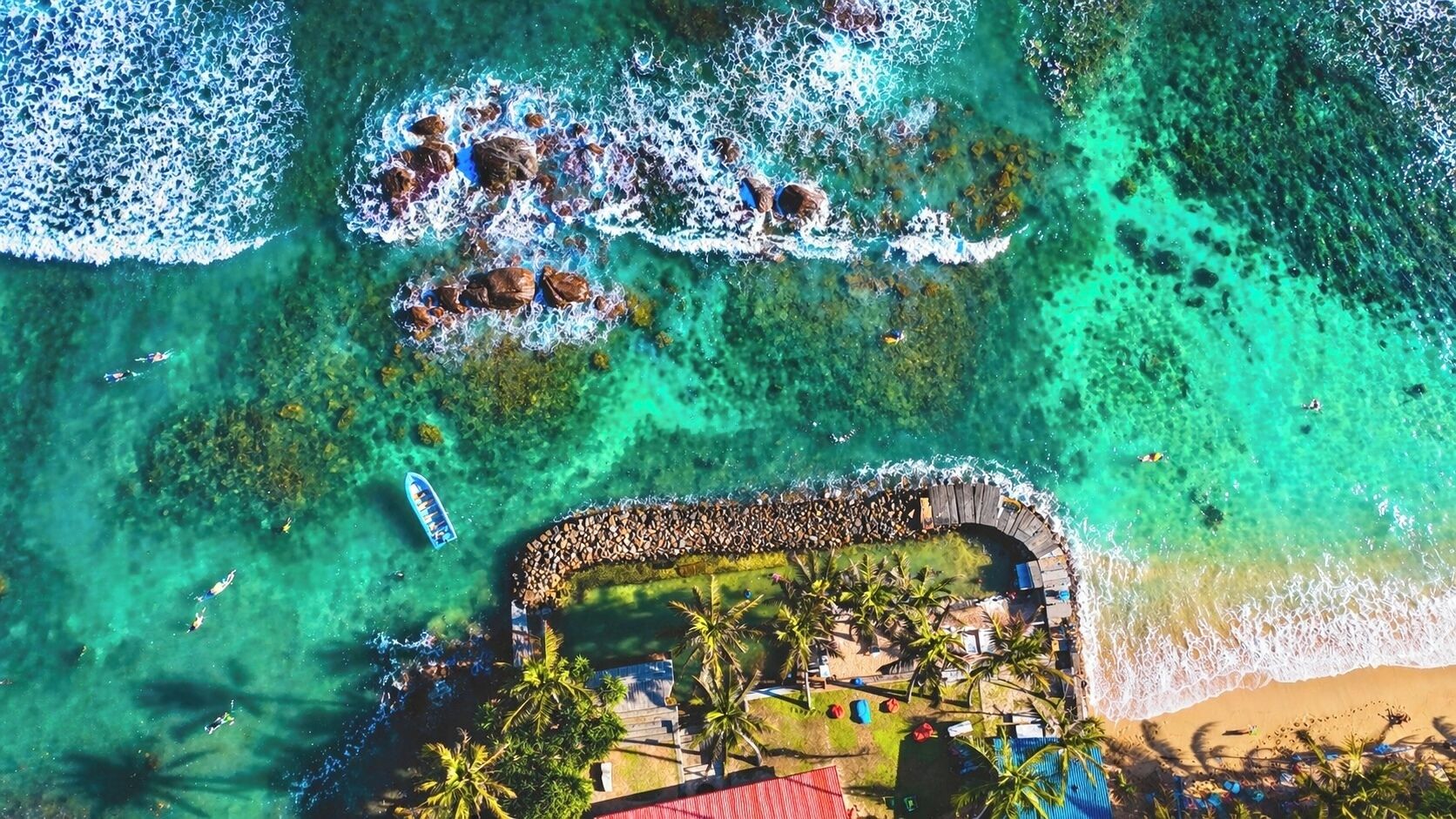 An aerial view of a beach resort with a wooden walkway, palm trees, and clear turquoise waters.