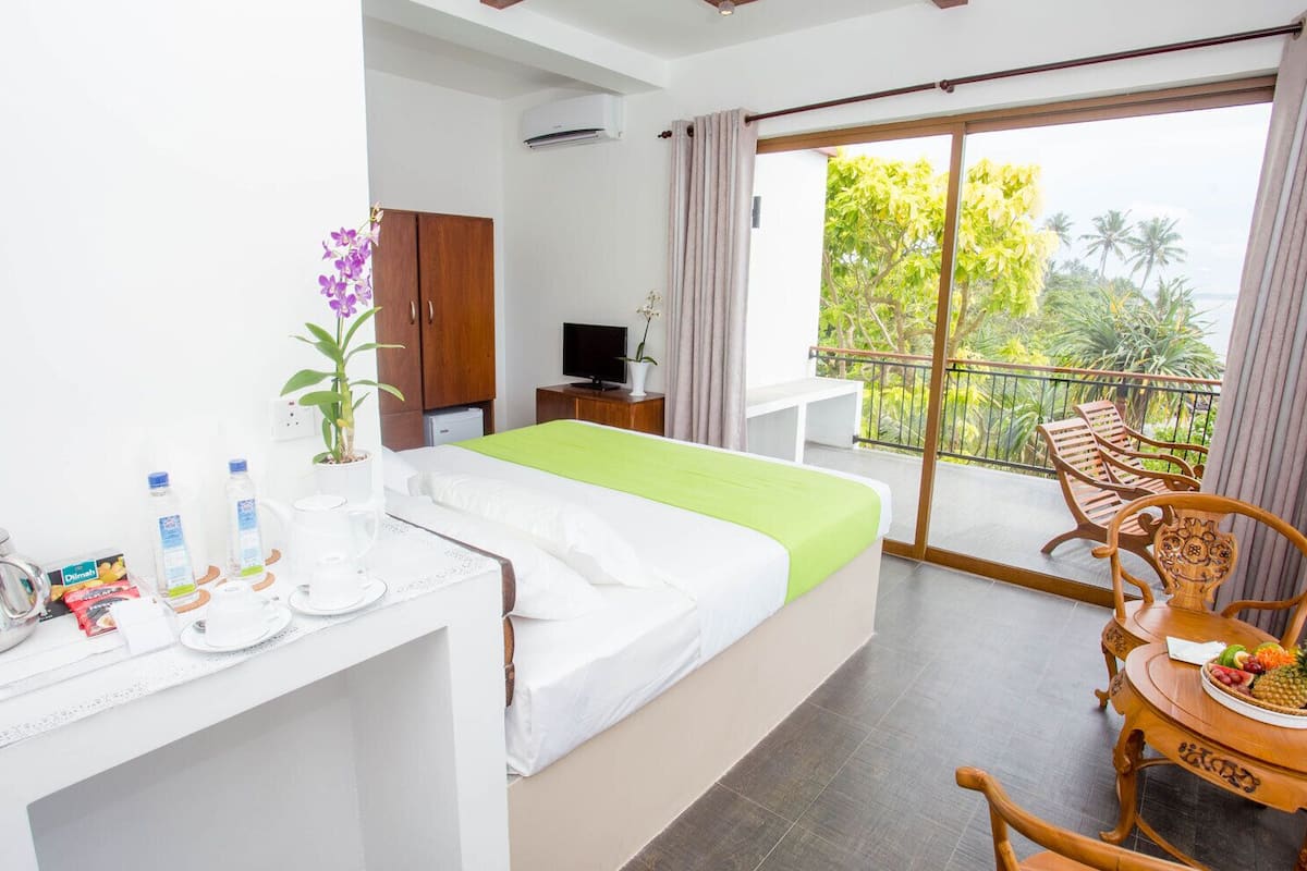 A hotel room with a bed, a desk with toiletries, a TV, and a balcony with chairs.