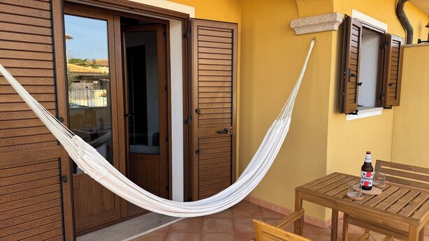 Una hamaca atada entre puertas de madera, una mesa con una botella y vasos, y una ventana con contraventanas.