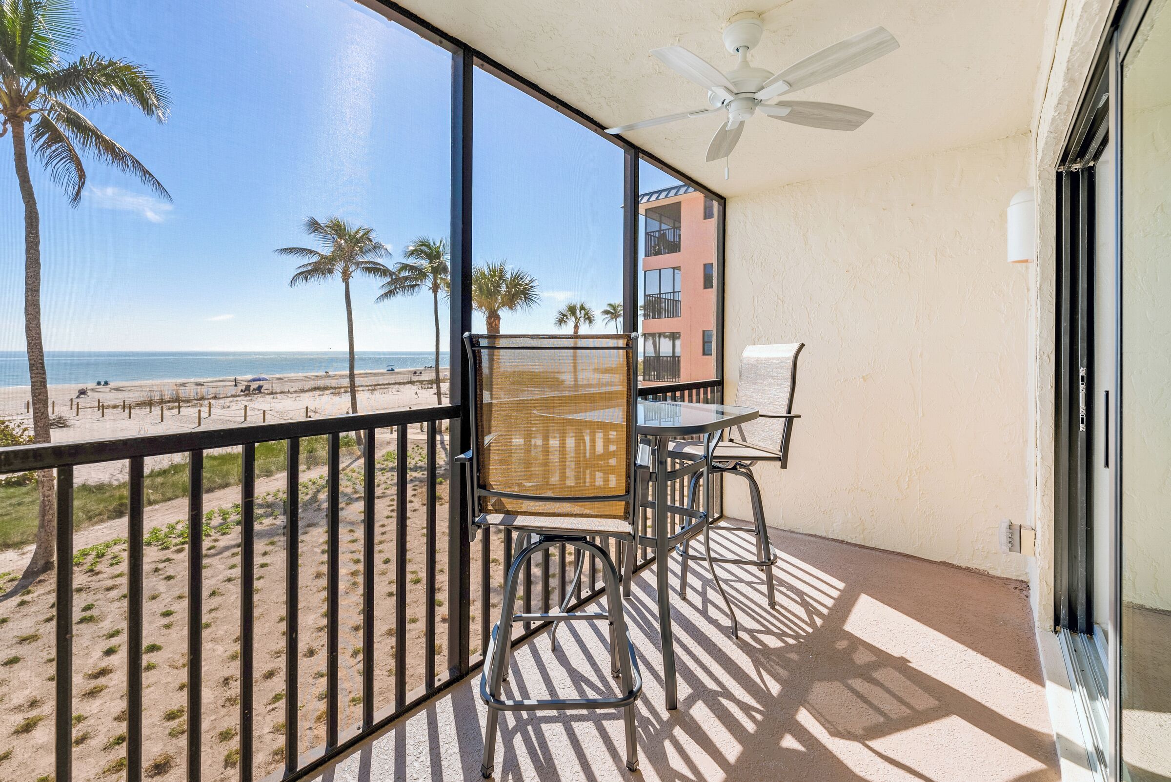 A balcony with a view of the beach, a ceiling fan, and a table with chairs.