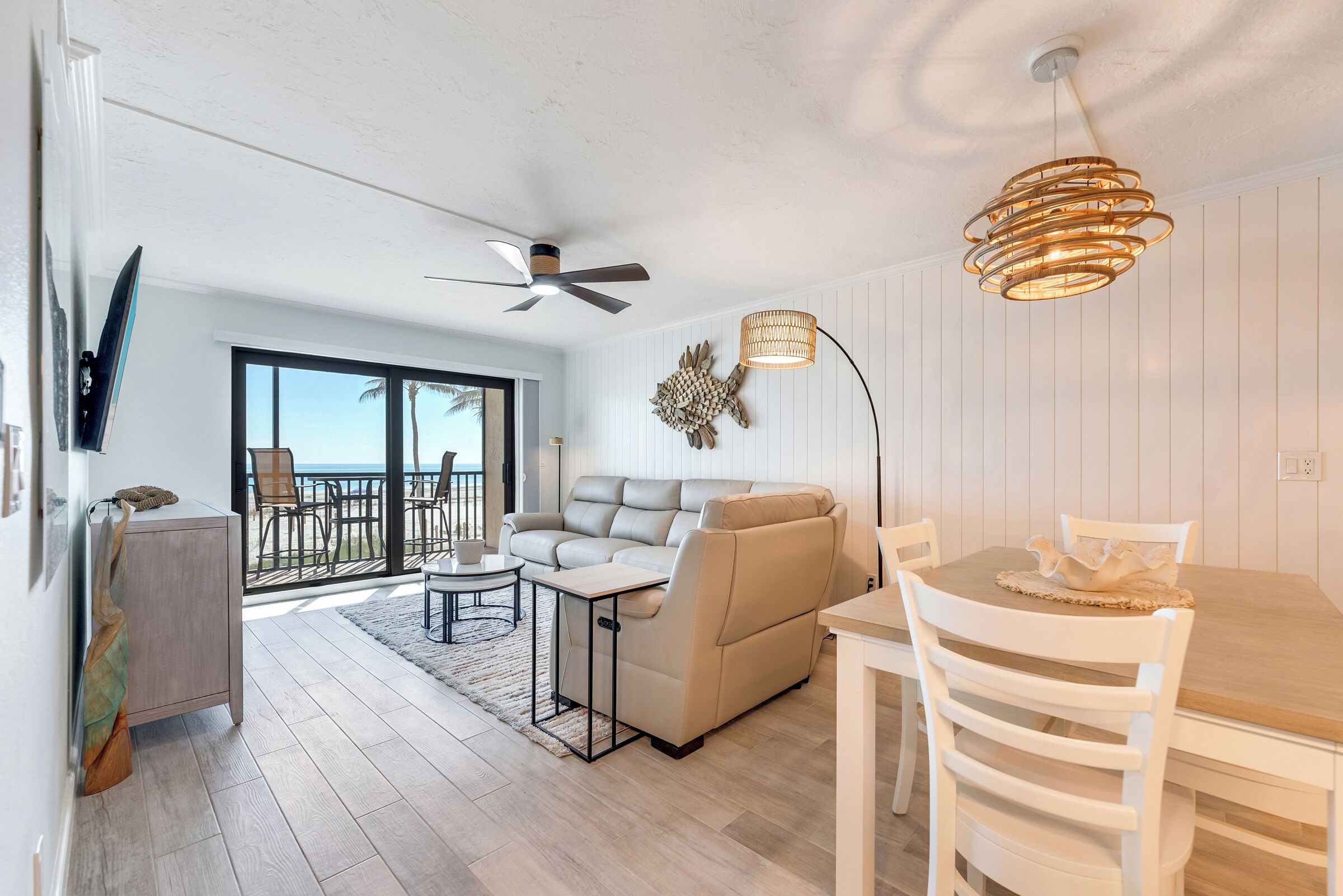 Una sala de estar moderna con sofá, mesa de comedor y ventilador de techo. Se ve el exterior a través de amplios ventanales.