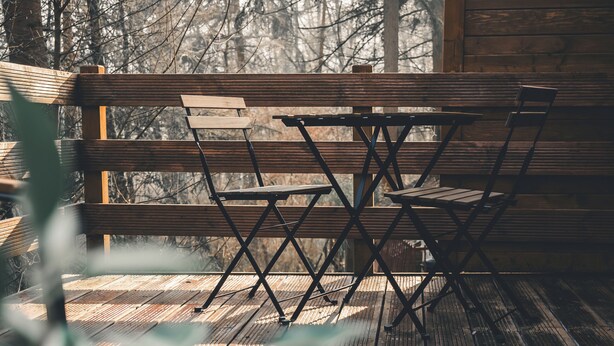 Een houten terras met een metalen klapstoel en een houten tafel, omringd door bomen.