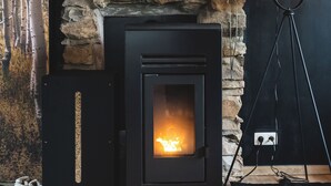 Een houtkachel met een achtergrond van een stenen muur, een vloerlamp en een paar sloffen op de vloer.