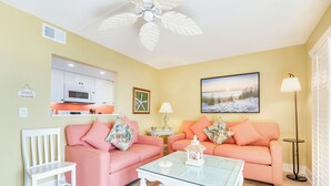 A living room with two pink sofas, a glass coffee table, a white chair, a kitchen area with white cabinets, a framed picture, and a floor lamp. - OCEAN'S REACH 2X2: 1-Bdrm, Gulf-Front View, All Amenities Open! (Sanibel Island)
