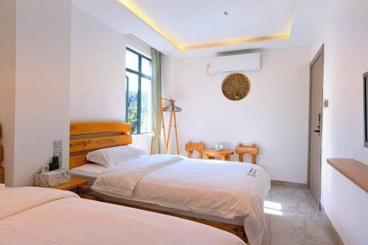 A hotel room with two beds, a wooden headboard, a small table with a lamp, a wall-mounted air conditioner, and a window with curtains.