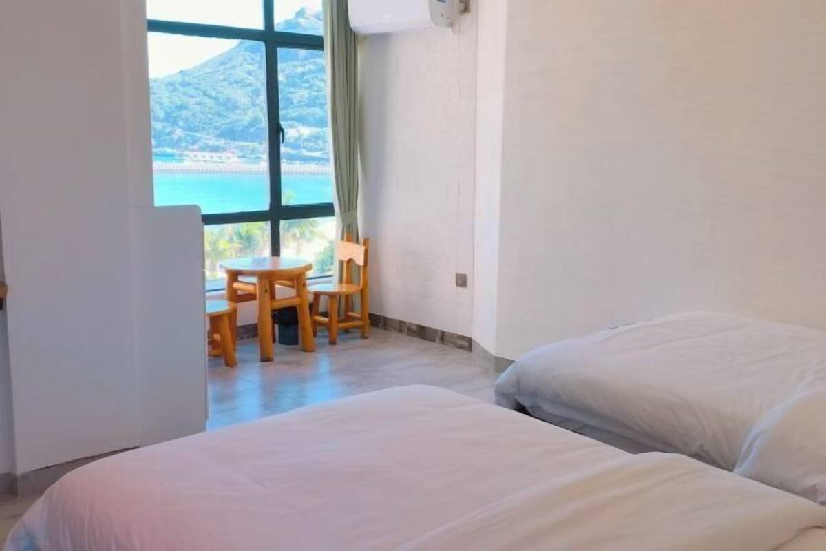 A hotel room with two beds, a small table with chairs, and a large window offering a view of a mountainous landscape.