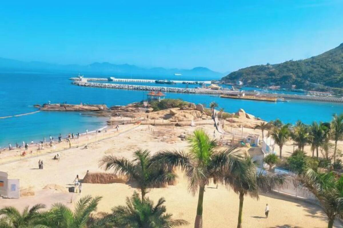 A beach with a pier, palm trees, and a mountainous backdrop.