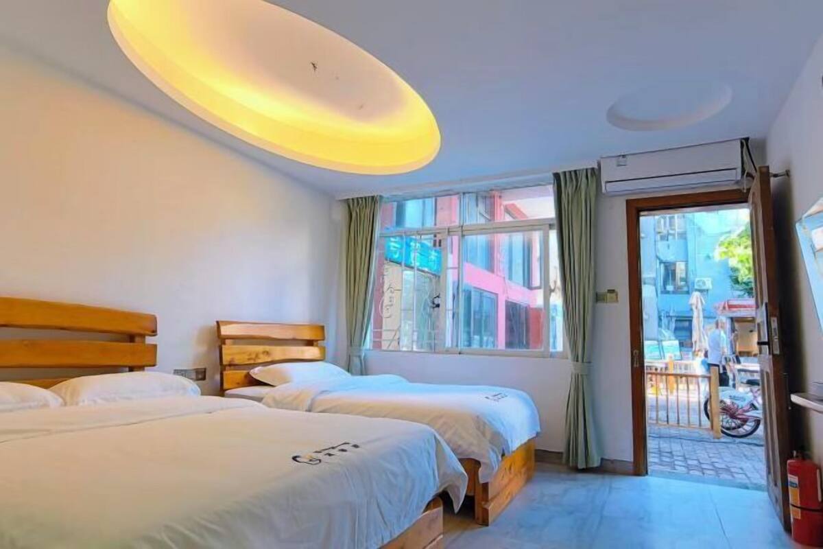 A hotel room with two beds, a window with curtains, an air conditioning unit, and a view of the street outside.