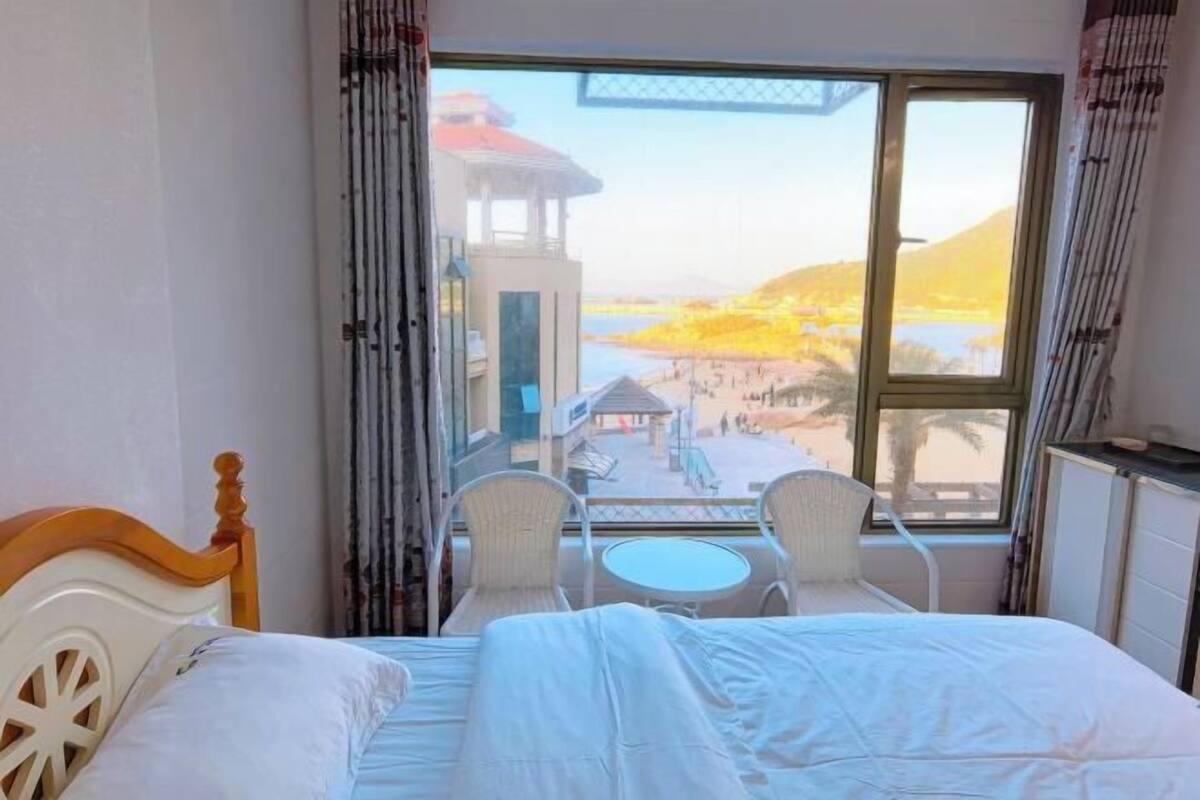 A hotel room with a bed, a small table, and two chairs. A large window offers a view of a beach and a building across the water.