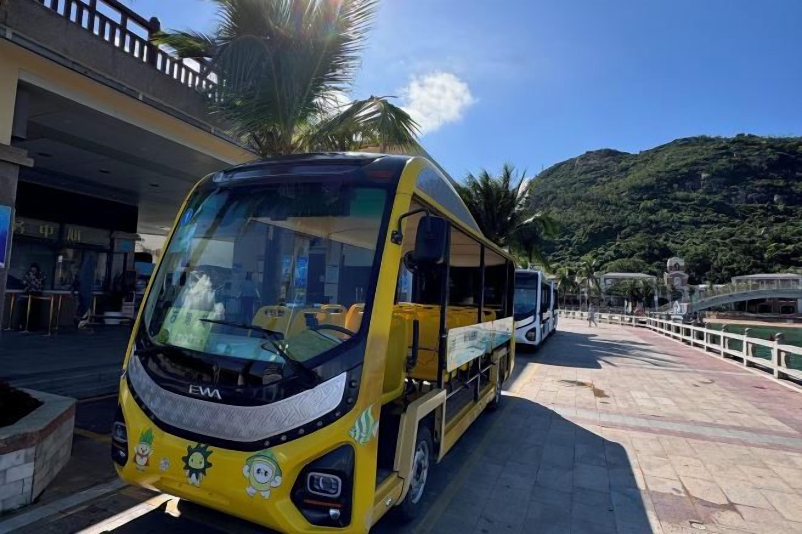 A yellow bus parked on a paved area with a bridge and greenery in the background.