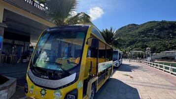 A yellow bus parked on a paved area with a bridge and greenery in the background.