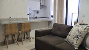 Un salon moderne avec un canapé et deux fauteuils en bois, un coin cuisine avec des meubles blancs et un plan de travail, et une grande fenêtre avec des rideaux.