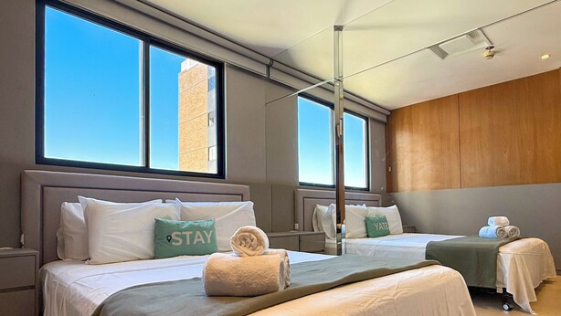 Une chambre d’hôtel avec deux lits, de grandes fenêtres, une vue sur un immeuble et un mur en bois.