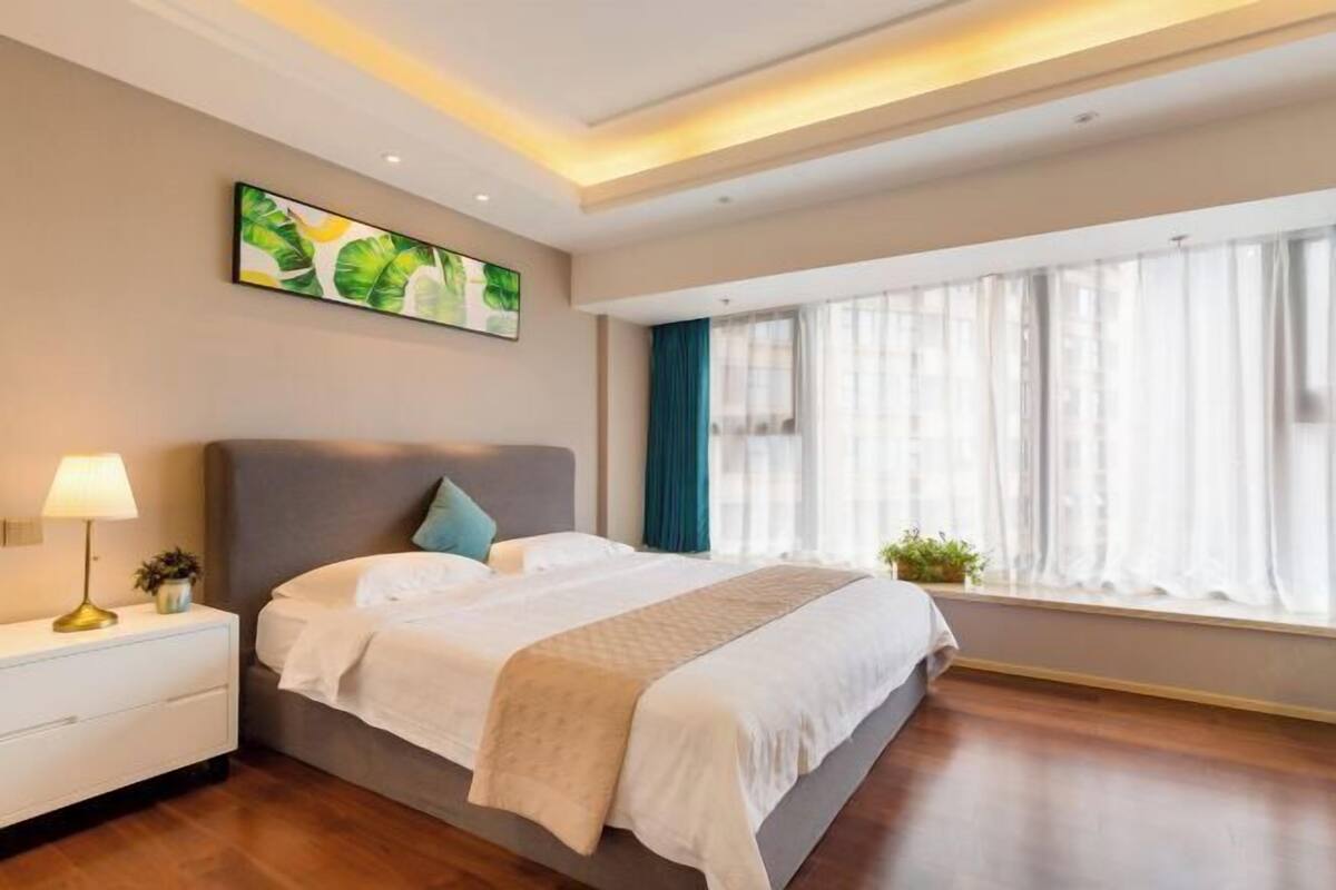 Ein modernes Hotelzimmer mit einem großen Bett, einem Nachttisch, einer Lampe, einem Bild an der Wand und einem Fenster mit Vorhängen.