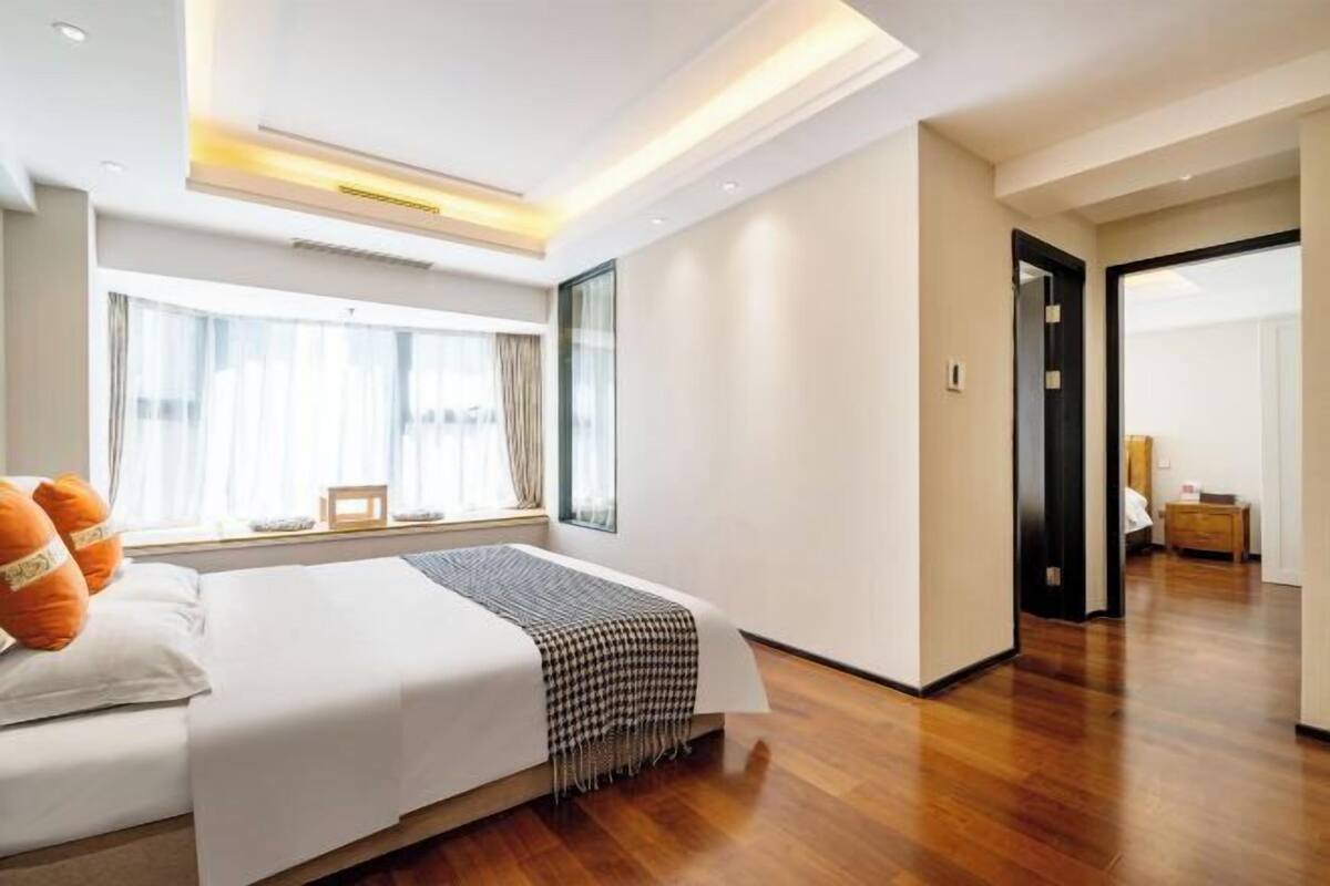 Ein modernes Hotelzimmer mit einem großen Bett, einem Fenster mit Vorhängen, einem Holzboden und einer Tür zu einem anderen Raum.