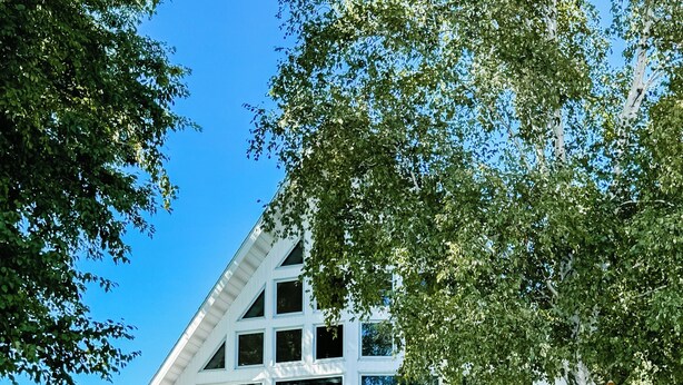 Un edificio blanco con techo inclinado, un muelle que da a una masa de agua y un área verde con mobiliario exterior.