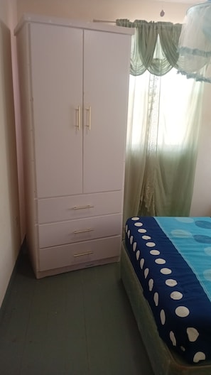 A bedroom with a large wardrobe, a bed with a blue and white polka dot bedspread, and sheer curtains.