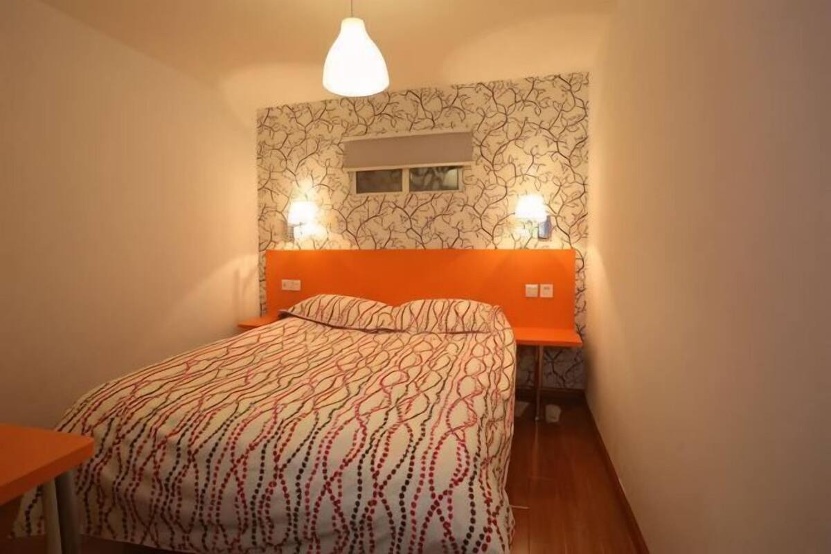 A bedroom with a patterned wall, a bed with an orange headboard, two bedside lamps, and a window.