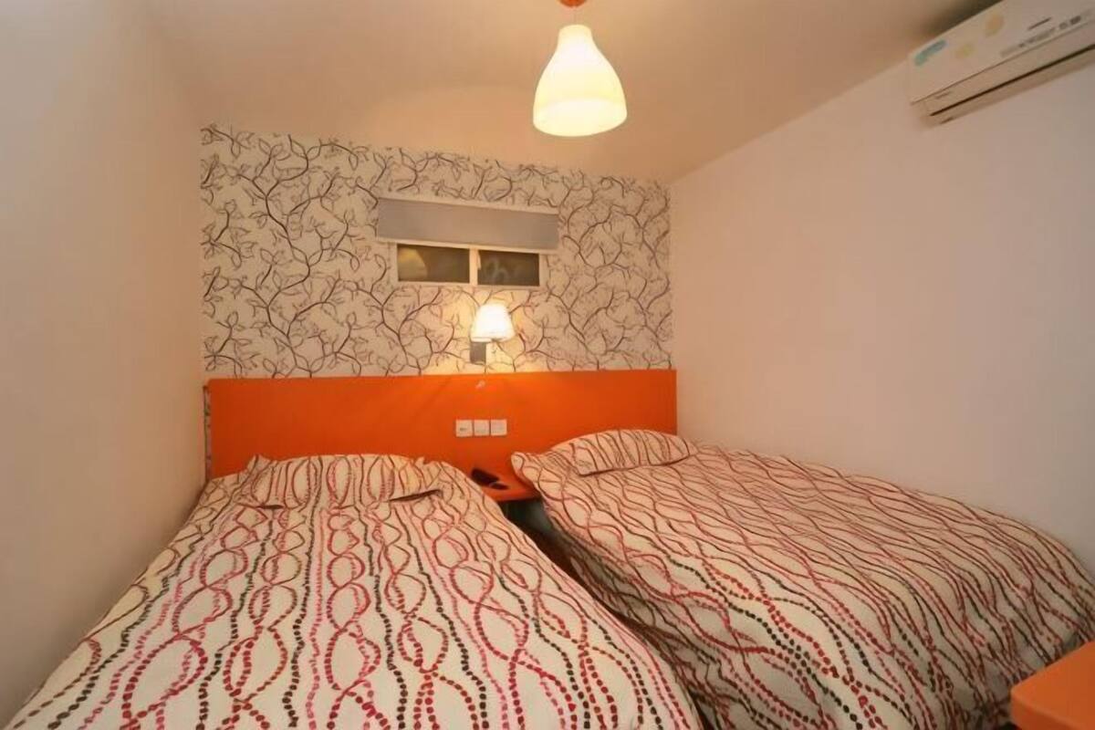 A room with two beds, a wall-mounted air conditioner, and a window with a light fixture.