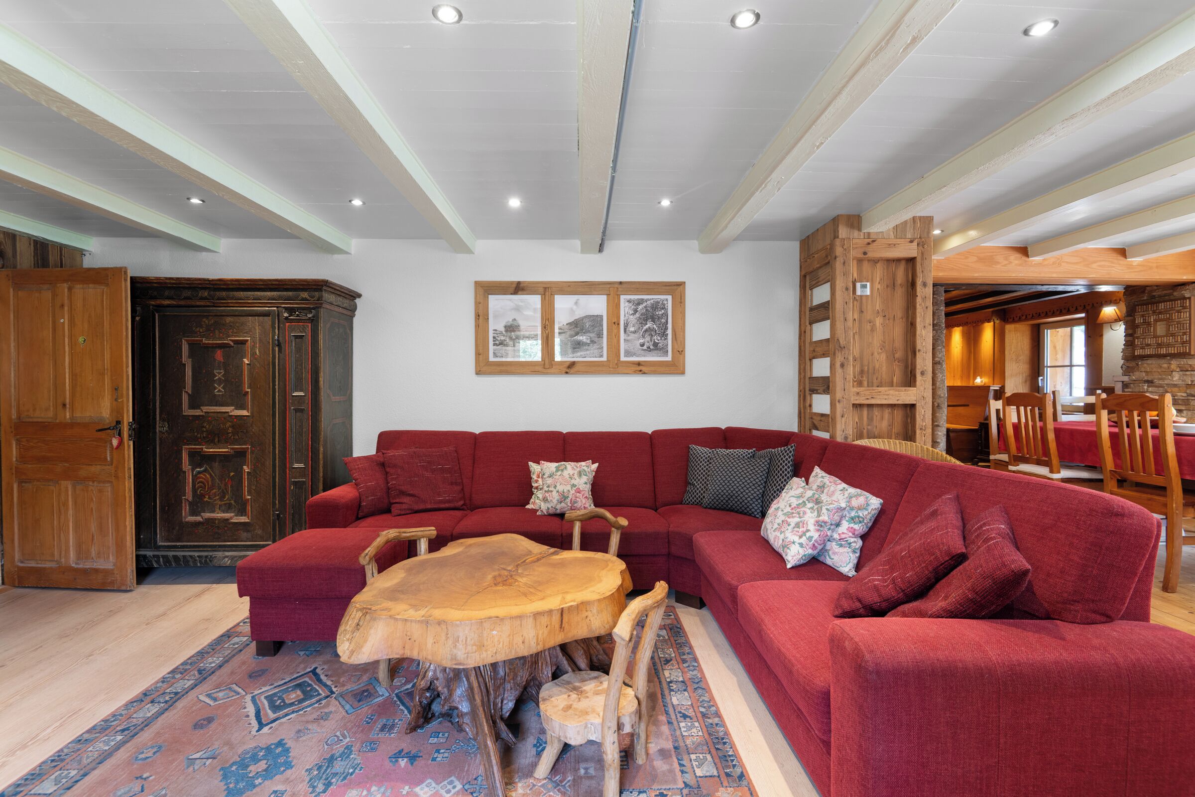 A red sofa, wooden dining table, and a patterned rug in a room with exposed wooden beams and a stone wall.