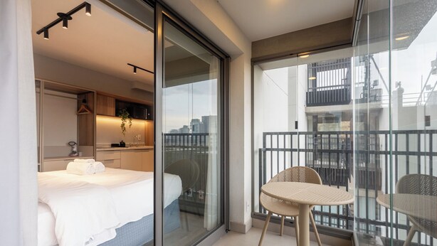 Una habitación de hotel moderna con cama, una kitchenette pequeña, un balcón con mesa y sillas, y vistas a la ciudad.